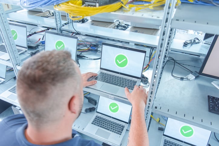 Ein IT-Mitarbeiter inspiziert auf Regalen gestapelte Laptops. Auf den Bildschirmen aller Laptops ist ein großes, grünes Häkchen zu sehen, das den erfolgreichen Abschluss der zertifizierten Datenlöschung signalisiert.