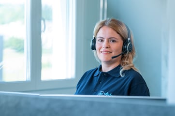 Lächelnende Mitarbeiterin eines IT-Service-Teams, die ein Headset trägt und an einem Schreibtisch sitzt.