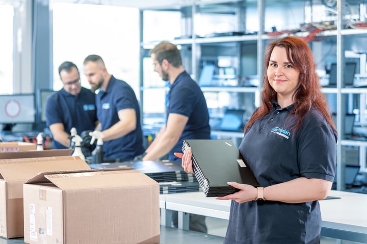 Eine dplan-Mitarbeiterin hält einen Stapel aufbereiteter Laptops in die Kamera, während im Hintergrund Kollegen in der Werkstatt IT-Hardware für das Remarketing prüfen und reinigen.