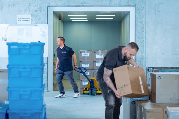 Zwei Mitarbeiter eines Grossunternehmens sind aktiv an einem IT-Massenrollout in der Schweiz beteiligt, dargestellt in einem Lager- oder Logistikbereich. Der Mann im Vordergrund, mit Tattoos am Arm, positioniert vorsichtig einen braunen Karton, vermutlich mit neuer IT-Hardware für die IT-Implementierung, auf einem Stapel weiterer Kartons, die teils das HP-Logo zeigen. Im Hintergrund zieht ein anderer Mann konzentriert einen blauen Hubwagen aus einem geöffneten Lastenaufzug oder einem Lagerzugang, beladen mit weiteren Lieferungen für den Massenrollout IT. Stapel von blauen Kunststoffbehältern im linken Vordergrund unterstreichen die logistische Komplexität des Projektmanagement Rollout. Ein Schild an der Wand weist auf eine maximale Bodenbelastung hin. Das Bild visualisiert die physische Arbeit und Koordination, die für die erfolgreiche IT Implementierung und den IT Rollout Schweiz unerlässlich ist.