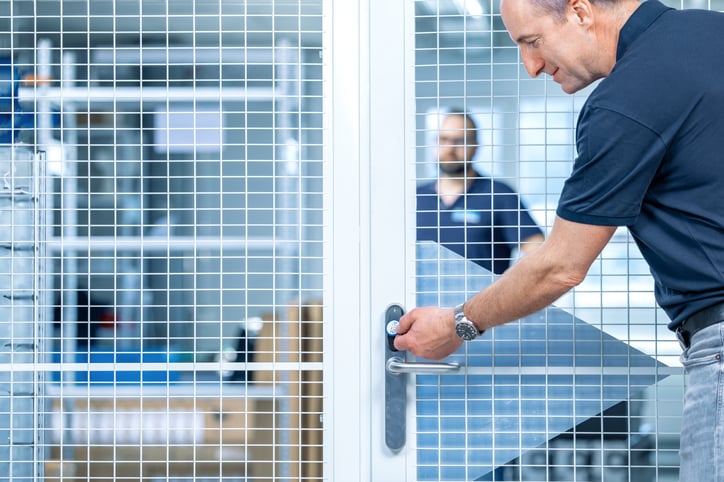 Ein Mann im dunkelblauen Polohemd öffnet mit einem elektronischen Schlüsselanhänger eine Gittertür in einem gesicherten Bereich, im Hintergrund steht eine weitere Person.